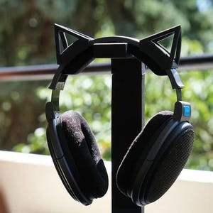 May include: Black over-ear headphones on a cat ear-shaped stand. The headphones have large, round earcups with soft, black padding. The stand is black and has a circular base. The cat ears are triangular.