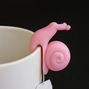 May include: A pink silicone snail tea bag holder perched on the rim of a white mug. The snail's body curls around the mug, with a tea bag hanging below. The background is black.