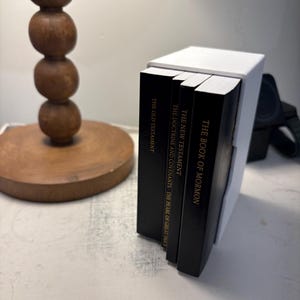 May include: A white book holder with four black books stacked inside. The books are titled "The Book of Mormon", "The New Testament", "The Doctrine and Covenants: The Pearl of Great Price", and "The Old Testament".