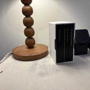May include: A white book holder with four black books. The books are titled "The Book of Mormon", "The New Testament", "The Doctrine and Covenants", and "The Pearl of Great Price". The book holder is on a white table next to a wooden lamp.