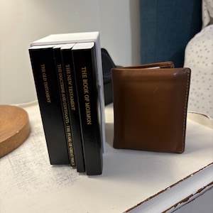 May include: A stack of four black hardcover books with gold lettering on a white surface. The books are titled "The Book of Mormon", "The New Testament", "The Doctrine and Covenants - The Pearl of Great Price", and "The Old Testament". A brown leather wallet is to the right of the books.