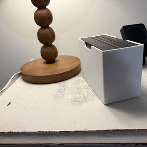May include: A white box with a black lid sits on a white table. The box is filled with black books. A wooden lamp with a round base is in the background.