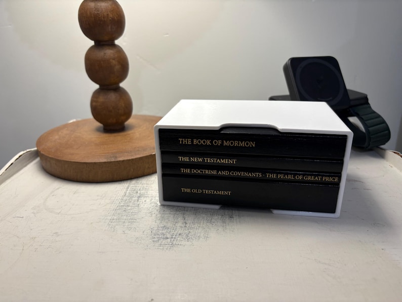 May include: A white box with black books stacked inside. The books are labeled "The Book of Mormon", "The New Testament", "The Doctrine and Covenants - The Pearl of Great Price", and "The Old Testament".
