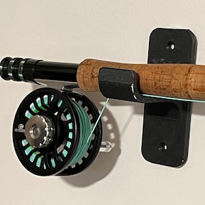 May include: A fly fishing rod and reel mounted on a black wall bracket. The rod has a cork handle and a black shaft. The reel is black with a green line. The bracket is mounted on a white wall.