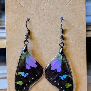 Peut inclure: Une paire de boucles d'oreilles en forme d'ailes de papillon avec des accents noirs, violets, verts et bleus. Les ailes sont enfermées dans de la résine transparente et pendent à des crochets argentés.