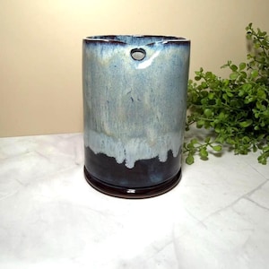May include: A cylindrical ceramic utensil holder featuring a reactive glaze with a dark brown base that transitions into a light blue and white drip pattern. The top rim is dark brown, and a small circular hole is positioned near the upper edge.