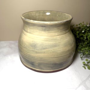 May include: A wide-mouthed ceramic vessel with a rounded body and a flared rim. The exterior features a cream-colored glaze with horizontal, dark gray, brush-like streaks that wrap around the circumference, creating a textured, layered appearance.