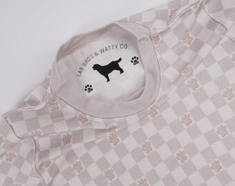 Könnte beinhalten: Ein hellgraues T-Shirt mit einem karierten Muster und Pfotenabdrücken. Das Shirt hat ein weißes Etikett mit dem Text "LAB BROS & WATTY CO." und einer Silhouette eines schwarzen Labrador Retrievers.