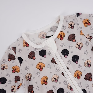 May include: A white baby onesie with a grey background and a pattern of brown, black, and yellow dogs with white paw prints. The onesie has a white zipper down the front.