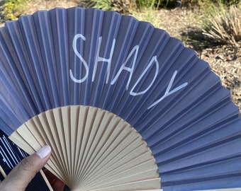 Może przedstawiać: Ręczny wachlarz z plisowanym, perwinkle-kolorowym papierowym wzorem. Słowo "SHADY" jest napisane na wachlarzu na biało. Uchwyt wachlarza jest wykonany z jasnego drewna.