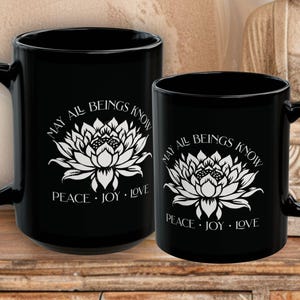 May include: Two black ceramic mugs with white floral designs and the text "MAY ALL BEINGS KNOW PEACE &bull; JOY &bull; LOVE". The mugs are different sizes, with a larger mug on the left and a smaller one on the right.