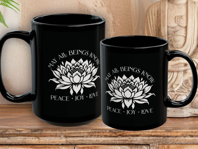 May include: Two black ceramic mugs with white floral designs and the text "MAY ALL BEINGS KNOW PEACE &bull; JOY &bull; LOVE". The mugs are different sizes, with a larger mug on the left and a smaller one on the right.