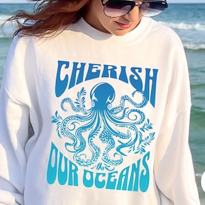 May include: White sweatshirt featuring a blue octopus design and the text "CHERISH OUR OCEANS". The octopus is surrounded by decorative elements. The sweatshirt is a casual style.
