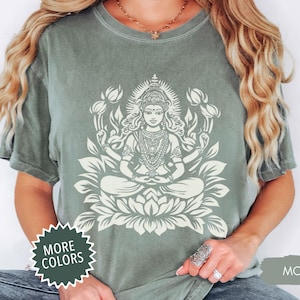 May include: Moss green t-shirt featuring a white graphic of a seated deity surrounded by a lotus flower and other floral elements. The shirt has short sleeves and a crew neck. The words "MORE COLORS" are in a white starburst.