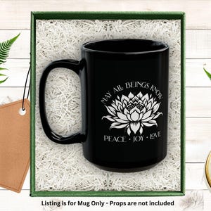 May include: Black ceramic mug with a white lotus flower design and the words "MAY ALL BEINGS KNOW PEACE JOY LOVE." The mug is presented in a green box with white filler and a blank tag. Fern leaves and a candle are in the background.