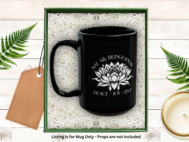 May include: Black ceramic mug with a white lotus flower design and the words "MAY ALL BEINGS KNOW PEACE JOY LOVE." The mug is presented in a green box with white filler and a blank tag. Fern leaves and a candle are in the background.
