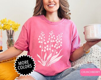 May include: A pink t-shirt with a white graphic of lily of the valley flowers. The text "MORE COLORS" is on a black starburst sticker. The text "CRUNCHBERRY" is on a pink sticker.