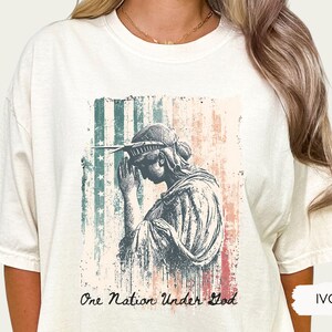 May include: Ivory-colored t-shirt featuring a distressed graphic of the Statue of Liberty with a faded American flag background. The text "One Nation Under God" is printed below the image. The shirt is a comfortable, casual style.
