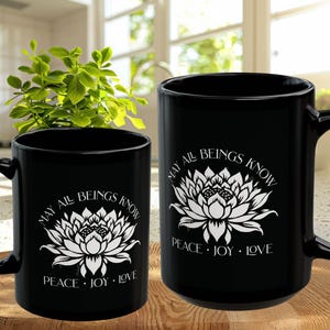 May include: Two black ceramic mugs with white floral designs and the text "MAY ALL BEINGS KNOW PEACE &bull; JOY &bull; LOVE". The mugs are on a wooden surface, with a green plant in the background.