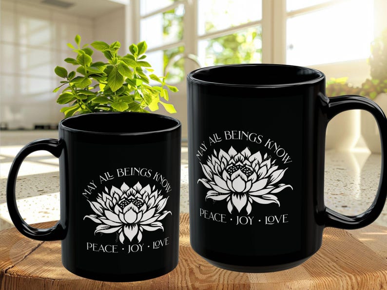 May include: Two black ceramic mugs with white floral designs and the text "MAY ALL BEINGS KNOW PEACE &bull; JOY &bull; LOVE". The mugs are on a wooden surface, with a green plant in the background.