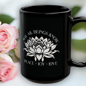 May include: Black ceramic mug with a white lotus flower design and the words "MAY ALL BEINGS KNOW PEACE &bull; JOY &bull; LOVE." Pink roses are in the background. The mug is approximately 5 inches tall.