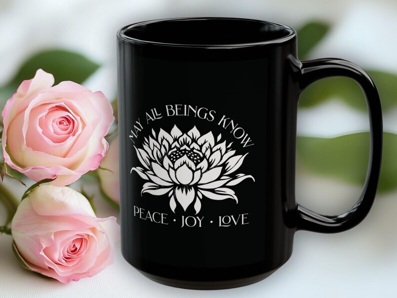 May include: Black ceramic mug with a white lotus flower design and the words "MAY ALL BEINGS KNOW PEACE &bull; JOY &bull; LOVE." Pink roses are in the background. The mug is approximately 5 inches tall.