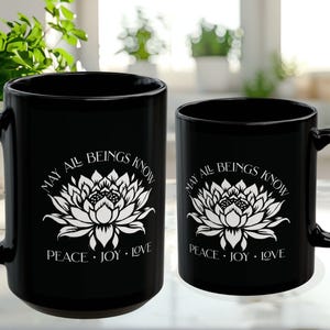 May include: Two black ceramic mugs of different sizes. Each mug features a white lotus flower design with the text "MAY ALL BEINGS KNOW PEACE &bull; JOY &bull; LOVE". The mugs have a curved handle.