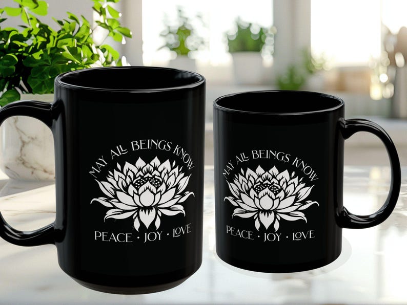May include: Two black ceramic mugs of different sizes. Each mug features a white lotus flower design with the text "MAY ALL BEINGS KNOW PEACE &bull; JOY &bull; LOVE". The mugs have a curved handle.