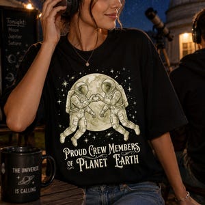 May include: Black t-shirt with a graphic of two astronauts forming a heart in front of a moon, surrounded by stars. The text "Proud Crew Members of Planet Earth" is below the graphic. A black mug with the text "The Universe is Calling" is on a wooden surface.