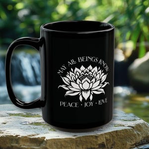 May include: Black ceramic mug with a white lotus flower design and the words "MAY ALL BEINGS KNOW PEACE &bull; JOY &bull; LOVE". The mug has a large handle and is set against a blurred natural background.