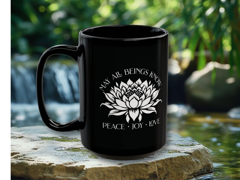 May include: Black ceramic mug with a white lotus flower design and the words "MAY ALL BEINGS KNOW PEACE &bull; JOY &bull; LOVE". The mug has a large handle and is set against a blurred natural background.