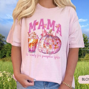 May include: Pink t-shirt with the text "MAMA is ready for pumpkin spice" in pink letters. The design features a watercolor pumpkin with a pink bow and a pink and orange iced coffee.