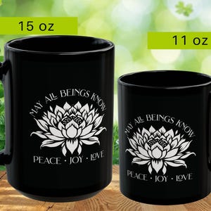 May include: Two black ceramic mugs, one 15 oz and the other 11 oz, with white floral designs and the text "MAY ALL BEINGS KNOW PEACE JOY LOVE". The mugs have large handles and are set against a blurred green background.