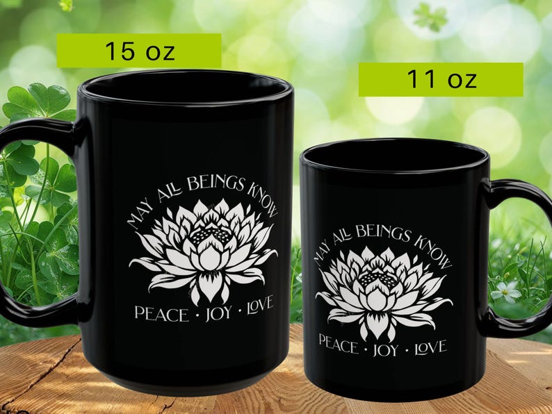 May include: Two black ceramic mugs, one 15 oz and the other 11 oz, with white floral designs and the text "MAY ALL BEINGS KNOW PEACE JOY LOVE". The mugs have large handles and are set against a blurred green background.