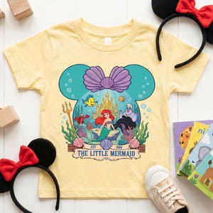 Puede incluir: Una camiseta amarilla clara con un gr&aacute;fico de La Sirenita. El dise&ntilde;o tiene forma de cabeza de rat&oacute;n con un lazo morado. El gr&aacute;fico incluye a Ariel, Flounder, Sebasti&aacute;n, &Uacute;rsula y un castillo. Se muestra el texto "The Little Mermaid".