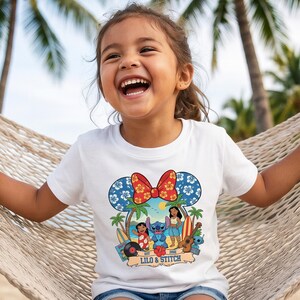 May include: White t-shirt featuring a colorful graphic of Lilo & Stitch. The design includes the characters Lilo, Stitch, and Nani, along with palm trees, surfboards, and the text "Lilo & Stitch Est 2001". The shirt has a Minnie Mouse ear design.
