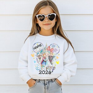 May include: A white sweatshirt featuring a Disney design with the text "Disney 2026". The design includes images of characters and a castle. The sweatshirt is worn by a person wearing heart-shaped sunglasses.