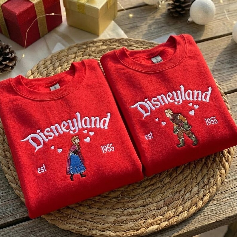 Puede incluir: Dos sudaderas rojas con la palabra "Disneyland" bordada en blanco. Cada una presenta un personaje y el a&ntilde;o "1955". Est&aacute;n sobre una estera tejida.
