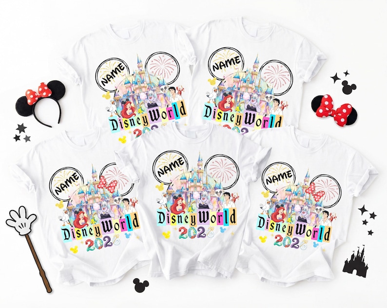 Puede incluir: Camisetas blancas con un dise&ntilde;o colorido de Disney World con un castillo, personajes y el texto "Disney World 2026". Las camisetas tienen un dise&ntilde;o de orejas de Mickey Mouse con la palabra "NAME" y fuegos artificiales. Accesorios: orejas de Minnie y una varita de guante de Mickey.