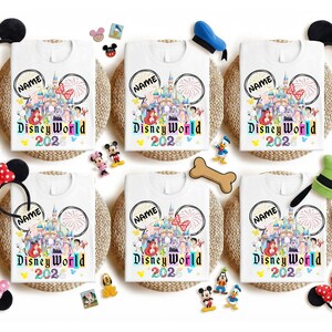 Puede incluir: Camisetas blancas con un dise&ntilde;o colorido de Disney World 2023, con un castillo, personajes y fuegos artificiales. Cada camiseta tiene un c&iacute;rculo "NAME" personalizable. Las camisetas est&aacute;n rodeadas de diademas con orejas de Mickey Mouse y figuras de personajes de Disney.