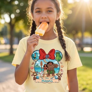 May include: A light yellow t-shirt featuring a colorful graphic of Moana characters. The design includes a red bow, ocean waves, and characters like Maui and Moana. The shirt is worn by a person eating a popsicle.