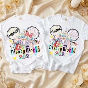 Puede incluir: Dos camisetas blancas con un dise&ntilde;o de Disney World. El dise&ntilde;o incluye un castillo, fuegos artificiales y personajes de Disney. El texto "Disney World 202" est&aacute; en letras coloridas, y hay un espacio para un nombre encima del castillo.