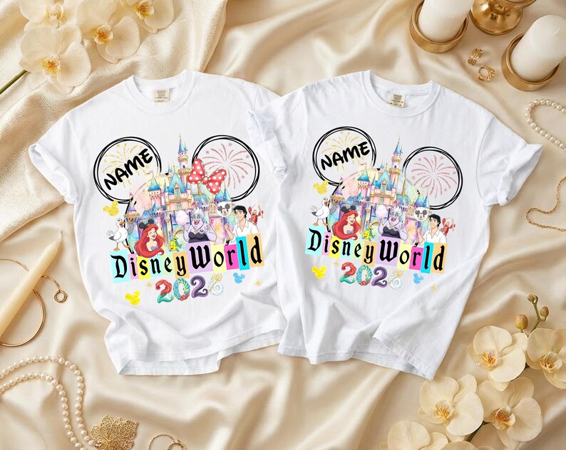 Puede incluir: Dos camisetas blancas con un dise&ntilde;o de Disney World. El dise&ntilde;o incluye un castillo, fuegos artificiales y personajes de Disney. El texto "Disney World 202" est&aacute; en letras coloridas, y hay un espacio para un nombre encima del castillo.
