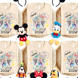 May include: Beige t-shirts with a Disney-themed graphic featuring characters and the text "NAME" and "2026". The shirts are arranged with Disney character plush toys and Mickey Mouse ears.