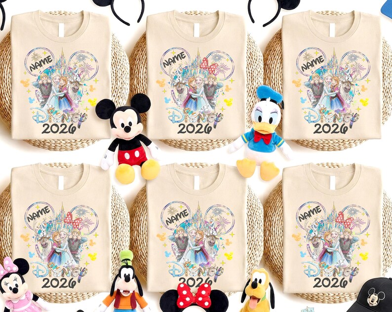May include: Beige t-shirts with a Disney-themed graphic featuring characters and the text "NAME" and "2026". The shirts are arranged with Disney character plush toys and Mickey Mouse ears.