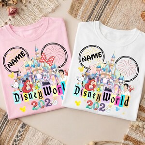 Puede incluir: Dos camisetas, una rosa y otra blanca, con un dise&ntilde;o de Disney World. El dise&ntilde;o incluye un castillo, personajes y las palabras "Disney World 2023". Cada camiseta tiene un contorno de oreja de Mickey Mouse negro y la palabra "NAME".