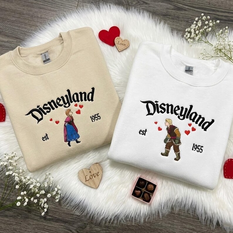 Puede incluir: Dos sudaderas, una beige y otra blanca, bordadas con "Disneyland" en letra cursiva. Cada una presenta una ilustraci&oacute;n de personaje, corazones y "est 1955". Tambi&eacute;n hay corazones decorativos y una caja de bombones.