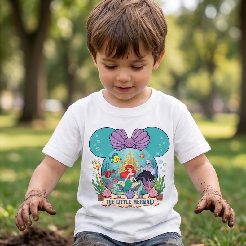Puede incluir: Camiseta blanca con un gr&aacute;fico colorido de La Sirenita de Disney. El dise&ntilde;o incluye a Ariel, Flounder, Sebasti&aacute;n y &Uacute;rsula, dentro de un marco con forma de Mickey Mouse. La camiseta tiene el texto "The Little Mermaid" y "Est. 1989".