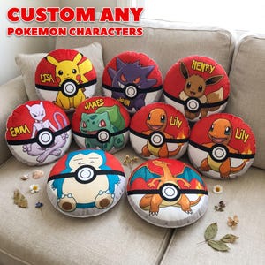 May include: A collection of round, red, white, and black Pokemon character pillows. Each pillow features a different character, such as Pikachu, Gengar, and Eevee, with a name printed on it. The pillows are arranged on a beige sofa.
