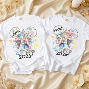 May include: Two white t-shirts featuring a colorful Disney design. The design includes the text "NAME" above a castle and characters from the movie Frozen, with the year "2026" below. One shirt has a red bow.